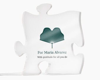 May include: A clear acrylic puzzle piece plaque featuring a dark green ginkgo leaf graphic. The text reads: For Maria Alvarez, With gratitude for all you do. The plaque is shaped like a standard jigsaw puzzle piece with smooth edges.
