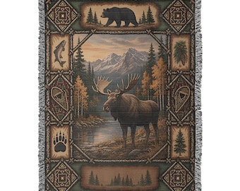 May include: A woven throw blanket featuring a central scene of a moose standing by a river with mountains and trees in the background. The border includes panels with a bear, a fish, pinecones, snowshoes, a paw print, and evergreen trees.