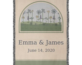 May include: A woven throw blanket in beige featuring a fringed edge and a central arch graphic containing a field of stylised flowers in blue, green, and yellow. The text "Emma & James" and "June 14, 2020" is printed in dark grey below the arch.