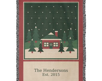 May include: A woven throw blanket featuring a winter scene with a red house, green pine trees, and white snowflakes on a dark green background. The bottom section is cream-coloured with the text The Hendersons Est. 2015 in dark grey.