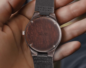 May include: A wristwatch with a dark brown wooden back casing and a dark grey leather strap. The back of the watch features the engraved text EMMA & JAMES JUNE 14, 2026. The circular wooden case is secured with four small silver-toned metal screws.
