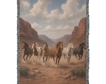 May include: A woven throw blanket featuring a scene of six horses galloping across a dusty desert landscape between tall red rock canyon walls under a blue sky with white clouds. The blanket has a decorative fringed edge on all sides.