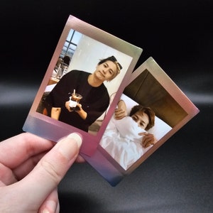 May include: Two instant photo prints with a rainbow border. The first photo shows a person wearing a black shirt and sunglasses holding a cup of coffee. The second photo shows a person lying in bed with a white blanket covering their face.