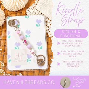 May include: A clear Kindle case with a floral design and a beaded strap. The strap features lavender and floral beads, along with a silver ring and a silver metal attachment. The text "Kindle Strap" is visible.