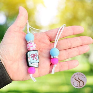 May include: Two pacifier clips with blue, pink, and white beads. One clip features a book charm with a pink bow. The other clip has a blue and pink bead design. Both clips have white cords.