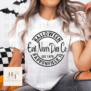 Könnte beinhalten: Weißes T-Shirt mit einem schwarzen, kreisförmigen Aufdruck mit der Aufschrift "HALLOWEEN Evil Never Dies Co. EST. 1978 HADDONFIELD, IL". Das Shirt hat einen Rundhalsausschnitt. Schwarze Fledermausdekorationen sind im Hintergrund zu sehen.