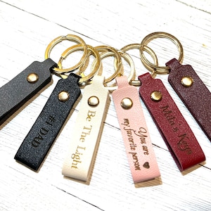 May include: Six leather keychains with gold key rings and engraved messages. The messages include "#1 Dad", "Be The Light", "You are my favorite person", "Mila's Keys", and two others.