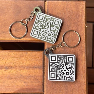 Könnte beinhalten: Zwei Schlüsselanhänger mit schwarzen und weißen QR-Codes auf einer Holzoberfläche. Der Schlüsselanhänger links ist grün und der rechts ist schwarz.