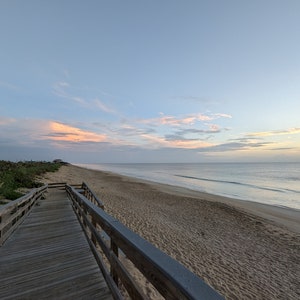 May include: A wooden boardwalk leads to a sandy beach with a blue sky and white clouds. The ocean is in the background with waves crashing on the shore.