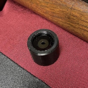 May include: A black cylindrical object with a central hole, possibly a component of a firearm or similar device. The object sits on a textured red surface, with a wooden element and a dark surface in the background.