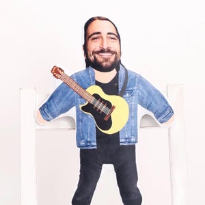 May include: A cardboard cutout of a smiling man with a beard, wearing a denim jacket and black pants, holding a yellow guitar with a black center. The cutout is propped up on a white bench.