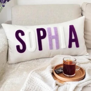 Puede incluir: Una almohada rectangular blanca con el nombre "SOPHIA" bordado en letras moradas, blancas y lavanda. La almohada está sobre un sofá de color claro. Una bandeja de madera con un vaso de té está delante.