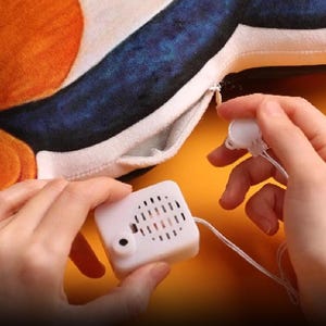 Op de afbeelding: Een pluche kussen met oranje en blauwe accenten, met een verborgen compartiment met een witte muziekdoos. De muziekdoos wordt vastgehouden door de handen van een persoon, met een trekkoord zichtbaar. Het ontwerp van het kussen is zichtbaar op de achtergrond.