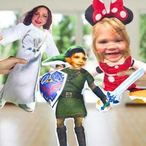 Op de afbeelding: Drie op maat gemaakte pluche poppen. Eén pop is gekleed in een witte jurk, een andere is gekleed als Link uit The Legend of Zelda, en de derde is gekleed als Minnie Mouse. Elke pop heeft een foto van het gezicht van een persoon.