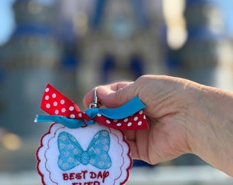 May include: A white, scalloped-edged keychain with a blue bow and the embroidered text "BEST DAD EVER." The keychain has a red and white polka dot ribbon and a blue ribbon. The background features a blurred castle.