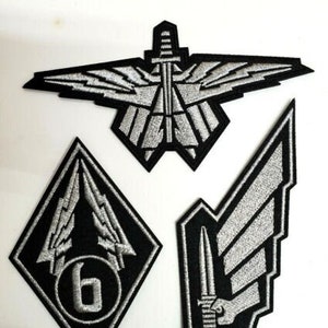 Op de afbeelding: Drie geborduurde patches in zwart en zilver. De eerste patch toont een gestileerde vogel met uitgespreide vleugels en een dolk in het midden. De tweede patch toont een gestileerde bliksemflits met het nummer "6" in het midden. De derde patch toont een gestileerde vleugel met een dolk in het midden.