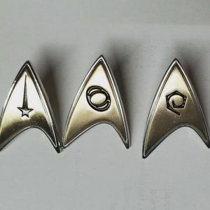 Pode incluir: Quatro distintivos metálicos do uniforme Star Trek. Cada distintivo é dourado com uma borda prateada e um símbolo único. Os símbolos incluem uma estrela, um círculo, uma forma estilizada e uma cruz vermelha.