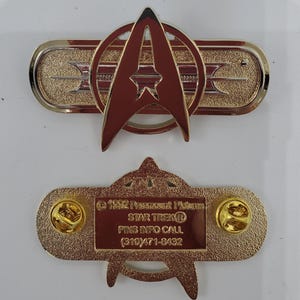 Puede incluir: Pin de Star Trek de color dorado con un emblema de la Flota Estelar rojo y blanco. La parte posterior del pin tiene la información de copyright, "1992 Paramount Pictures", "STAR TREK", "PINS INFO CALL (310) 471-8432".