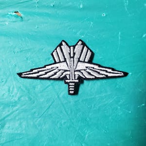 May include: Embroidered patch with a symmetrical design, featuring wings and a central vertical element. The patch is predominantly white with black edging, creating a detailed, textured appearance. The design resembles a military or organizational insignia.