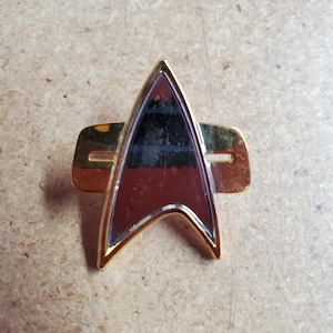 May include: A gold-colored Star Trek insignia pin with a red and black delta symbol. The pin has a gold-colored bar across the back.