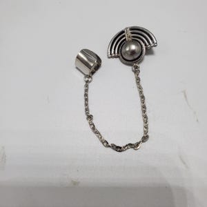 May include: Silver ring with a chain attached to a silver charm with a pearl-like center and a geometric design. The charm is shaped like a half-circle.