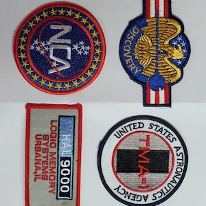 Könnte beinhalten: Vier gestickte Patches mit NASA-Logos. Der obere linke Patch ist blau mit weißen Sternen und dem Text "NCA". Der obere rechte Patch ist blau mit einem goldenen Adler und dem Text "DISCOVERY". Der untere linke Patch ist grau mit einem blauen Rechteck und dem Text "HAL 9000 LOGIC MEMORY SYSTEMS URBANA, IL". Der untere rechte Patch ist weiß mit einem grauen Rechteck und dem Text "TMA UNITED STATES ASTRONAUTICS AGENCY".