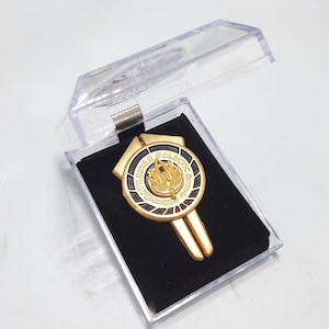 Puede incluir: Un pin de metal dorado con fondo negro. El pin presenta las palabras "Battlestar Galactica" y una imagen estilizada de un fénix.