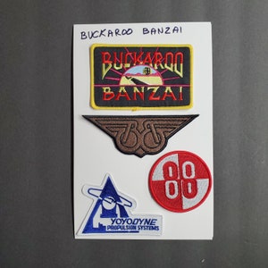 May include: Four embroidered patches on a white background. The top patch is black with yellow trim and reads "BUCKAROO BANZAI". The second patch is brown with a stylized "B&B" logo. The third patch is red and white with the number "88" in the center. The fourth patch is blue and white with a stylized rocket ship and the text "YOYODYNE PROPULSION SYSTEMS".