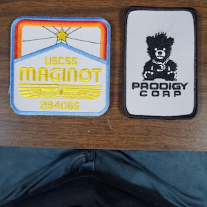 May include: Two embroidered patches. One is rectangular with a blue, orange, and yellow design and the text "USCSS MAGINOT" and "294065". The other is a gray rectangle with a black bear design and the text "PRODIGY CORP".
