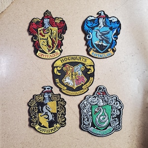 May include: Embroidered patches featuring the crests of Hogwarts houses: Gryffindor, Ravenclaw, Hufflepuff, and Slytherin, along with a Hogwarts crest. Each patch has detailed stitching and vibrant colors, with text identifying each house.