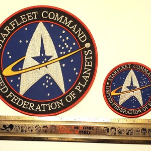 Może przedstawiać: Dwa haftowane naszywki z logo Starfleet Command. Większa naszywka ma około 10 cm średnicy, a mniejsza około 5 cm średnicy. Obie naszywki mają niebieskie tło z białym emblematem Starfleet i żółtym pierścieniem.