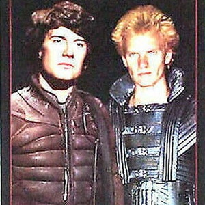 May include: A vintage movie poster for the film "DUNE" featuring two men in dark clothing. The poster has a red background with the title "DUNE" in white letters.