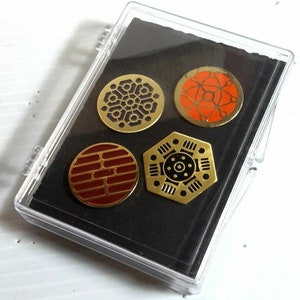Pode incluir: Um conjunto de quatro pinos de metal dourado com diferentes desenhos geométricos. Os pinos são exibidos em uma caixa de plástico transparente com fundo preto.