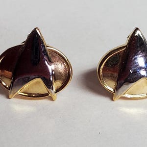 May include: A pair of gold-tone earrings with a silver-tone Starfleet insignia. The earrings are oval-shaped and have a smooth, polished finish.
