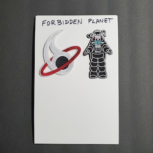 May include: Two iron-on patches on a white background. The left patch is a red, white, and blue stylized symbol with the text "FORBIDDEN PLANET". The right patch is a black silhouette of a robot.