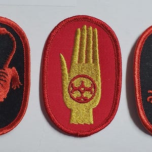 Peut inclure: Trois écussons brodés de forme ovale. Les écussons présentent un tigre rouge, une main jaune avec un symbole rouge et un dragon rouge, le tout sur des fonds noirs ou rouges. Les écussons ont des bordures rouges.