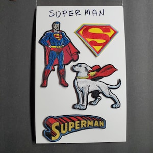 May include: Four embroidered patches featuring Superman. The patches include a red and yellow Superman logo, a red and yellow Superman logo with a white dog wearing a red cape, a red and yellow Superman logo with the word "SUPERMAN" in yellow letters, and a cartoon image of Superman in a blue and red suit.
