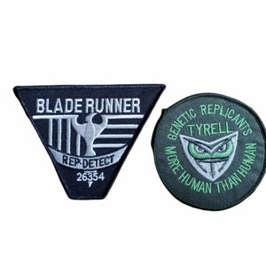 Puede incluir: Dos parches bordados negros y verdes. El primer parche es un triángulo con el texto "BLADE RUNNER" y "REP-DETECT 26354". El segundo parche es redondo con el texto "GENETIC REPLICANTS TYRELL MORE HUMAN THAN HUMAN".
