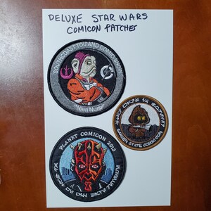 May include: Three embroidered Star Wars Comic Con patches. The first patch is gray with a cartoon of Nien Nunb from the movie Return of the Jedi. The second patch is brown with a cartoon of a Jawa from the movie Star Wars. The third patch is gray with a cartoon of Darth Maul from the movie Star Wars: Episode I - The Phantom Menace.
