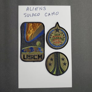 Könnte beinhalten: Drei gestickte Patches mit dem USCM-Logo, dem USS Sulaco-Logo und einem stilisierten Raumschiff-Design. Die Patches sind grün, schwarz und weiß.