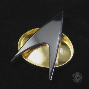 Puede incluir: Un emblema metálico de color plateado con forma de delta superpuesto sobre una base ovalada de color dorado. El emblema presenta un diseño afilado y puntiagudo con una superficie lisa y reflectante. El texto Quantum Mechanix es visible abajo.