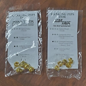 May include: Two plastic bags containing gold-coloured metal ranking pips for Star Trek characters. One bag is for Star Trek: Deep Space Nine, the other for The Next Generation. Each bag has a printed list of characters and their rank.