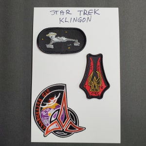 May include: Three embroidered patches with Star Trek Klingon designs. The top patch is an oval with a ship design. The middle patch is a red and black design. The bottom patch is a circular design with a purple and red emblem.