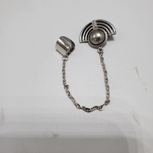 May include: Silver ring with a chain attached to a silver charm with a pearl-like center and a geometric design. The charm is shaped like a half-circle.
