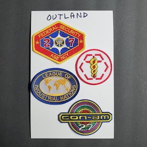 Peut inclure: Quatre patchs brodés avec des motifs et des couleurs différents. Les patchs sont sur un fond blanc. Les patchs sont pour une série télévisée de science-fiction fictive appelée "Outland". Les patchs comprennent les mots "Federal Security", "207", "Age Ncy", "League of Industrial Nations", "Con-am", et "27".