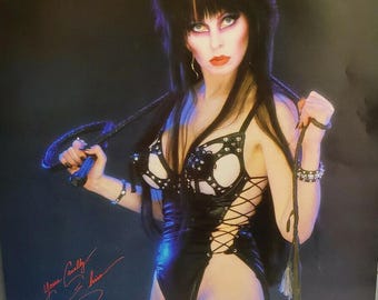 Può includere: Poster con una donna dai capelli scuri e trucco marcato, che indossa un completo in pelle nera con dettagli stringati. Tiene in mano una frusta e presenta una firma nell'angolo in basso a sinistra.