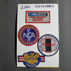 Peut inclure: Quatre patchs brodés, dont un patch NASA bleu et rouge avec le texte "NCA" et un patch blanc avec le texte "UNITED STATES ASTRONAUTICS AGENCY" et le texte "TMA-1" en rouge. Les patchs sont sur un fond blanc avec le texte "2001 DISCOVERY" en haut.
