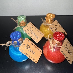 May include: A collection of four potion bottles with glittery contents in blue, green, gold, and red. Each bottle has a cork stopper and a tag with a handwritten label: "Mana", "Stamina", "Strength", and "Health". The bottles are tied with twine.