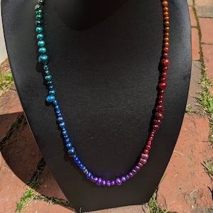 Pode incluir: Um colar degrad&ecirc; arco-&iacute;ris feito de p&eacute;rolas iridescentes. O colar &eacute; exibido em um busto de manequim preto.