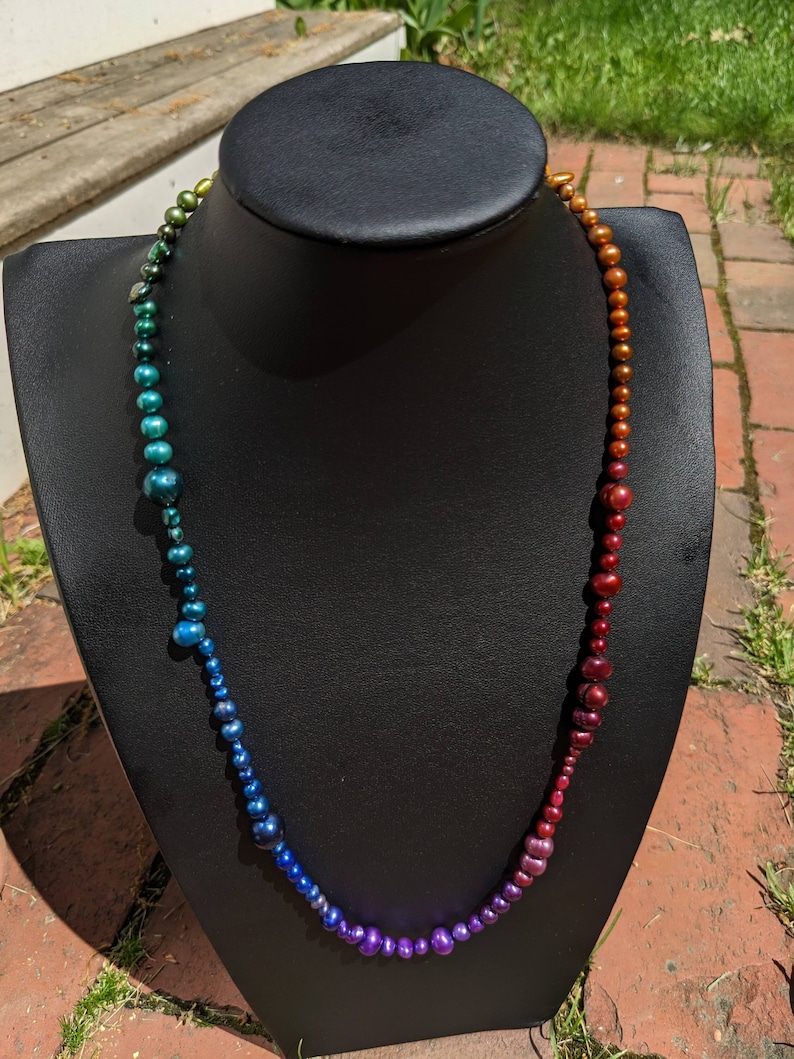 Pode incluir: Um colar degrad&ecirc; arco-&iacute;ris feito de p&eacute;rolas iridescentes. O colar &eacute; exibido em um busto de manequim preto.